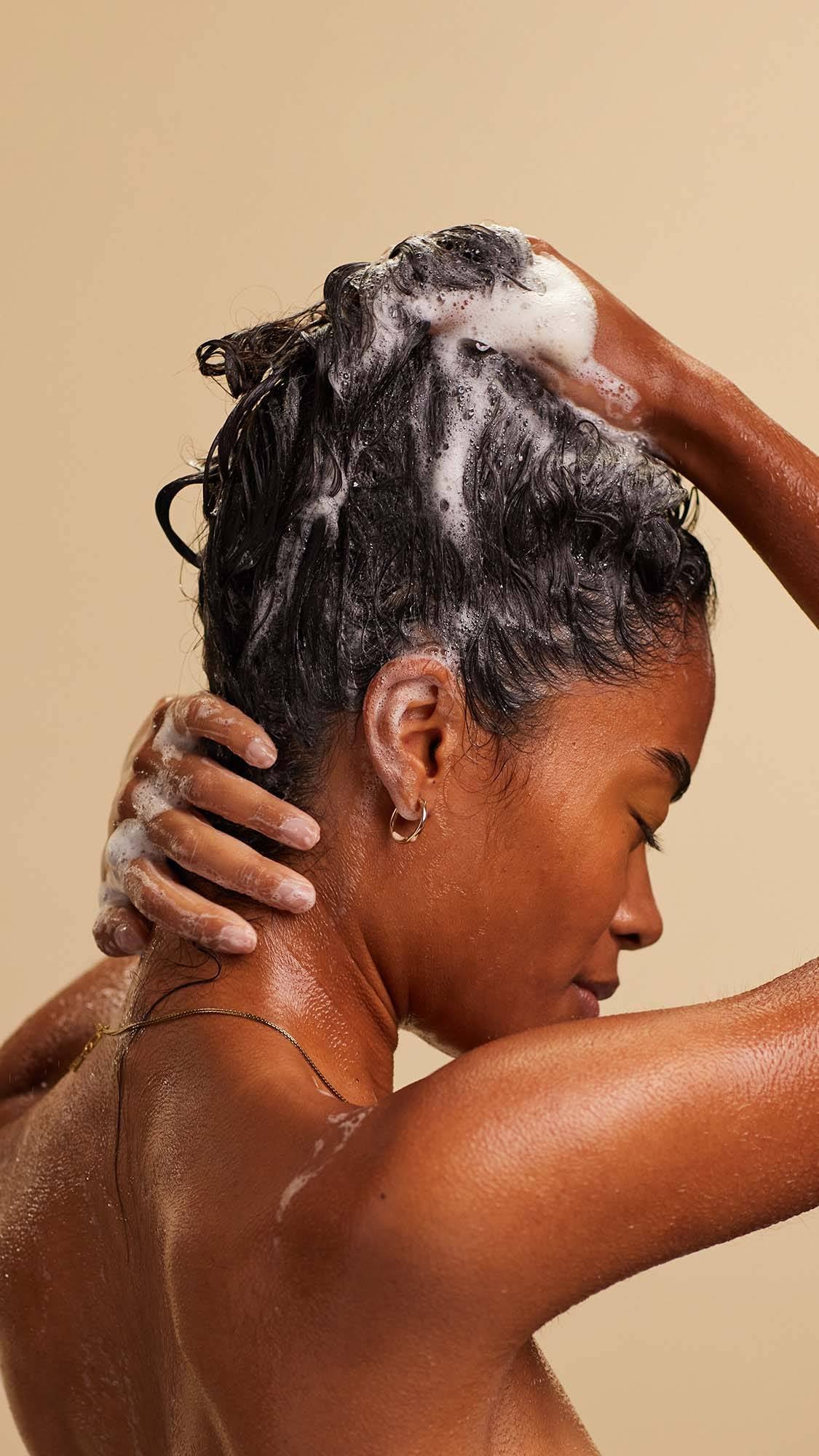 A blissed out, smiling person with long, dark, textured hair massages a white foaming shampoo into the lengths of their hair.