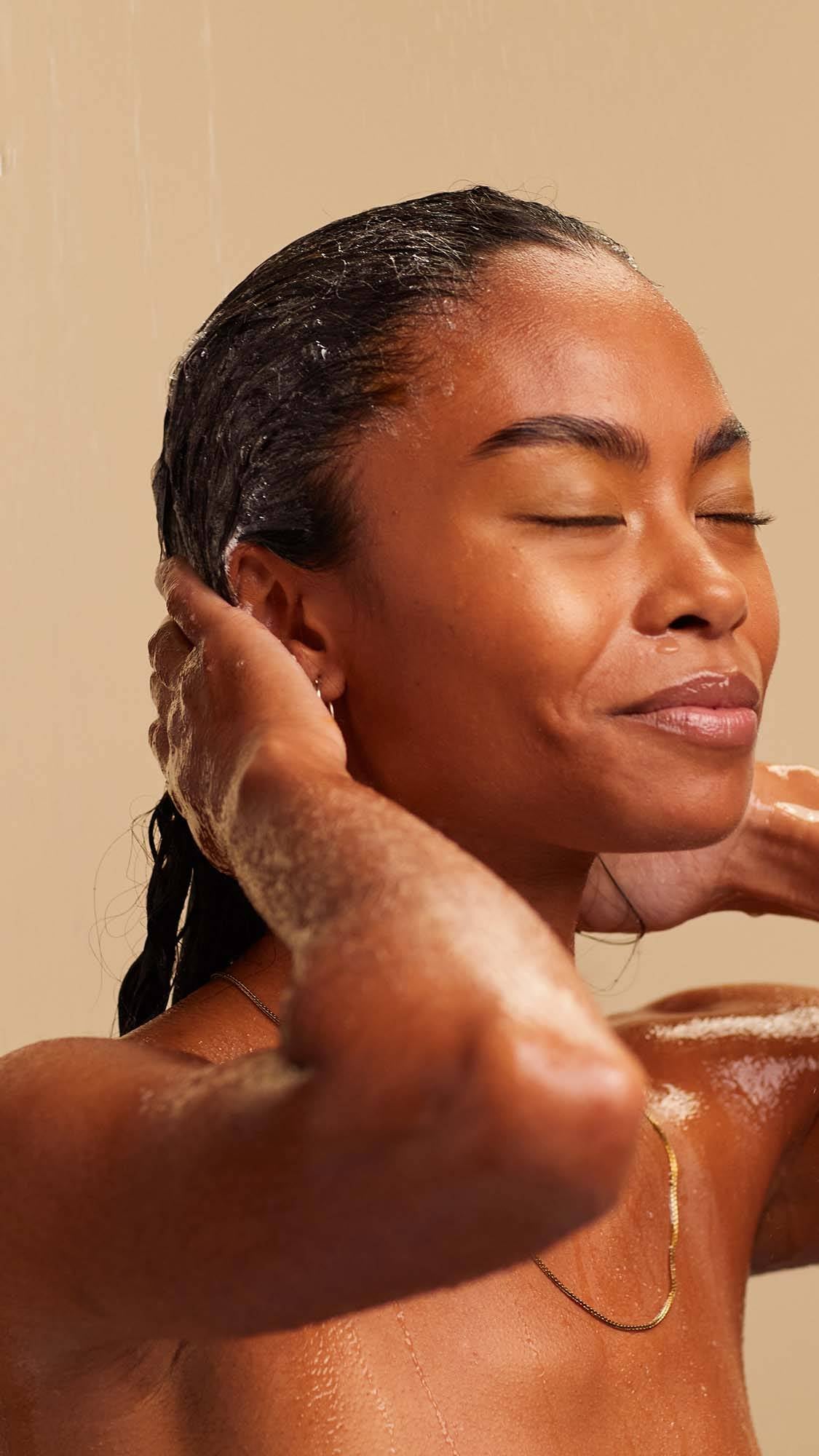 A blissed out, smiling person with long, dark, textured hair massages a white foaming shampoo into the lengths of their hair.