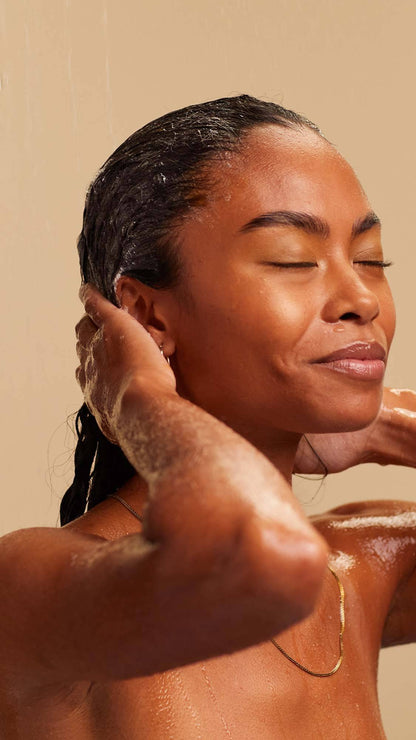 A blissed out, smiling person with long, dark, textured hair massages a white foaming shampoo into the lengths of their hair.