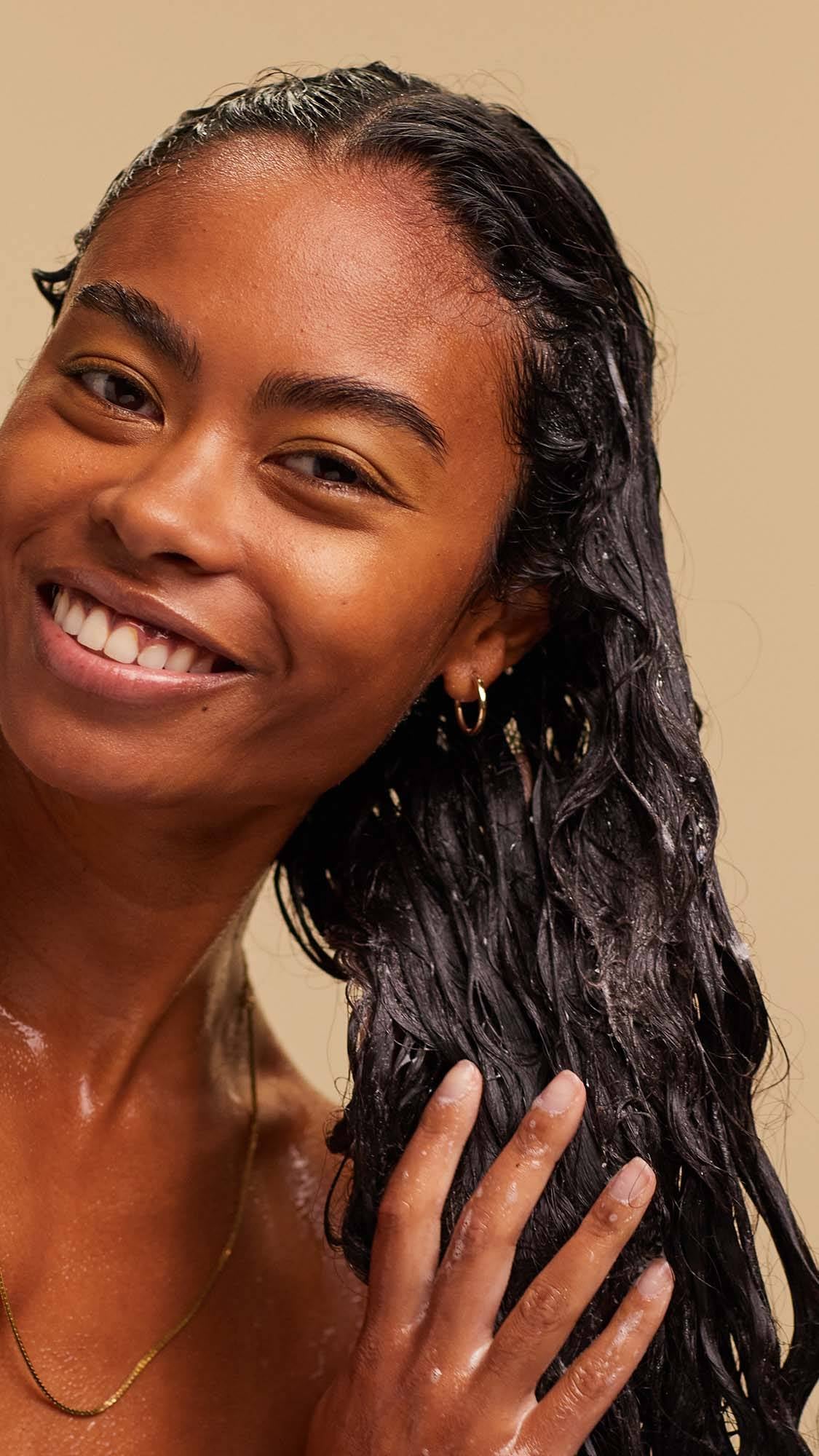 A blissed out, smiling person with long, dark, textured hair massages a white foaming shampoo into the lengths of their hair.