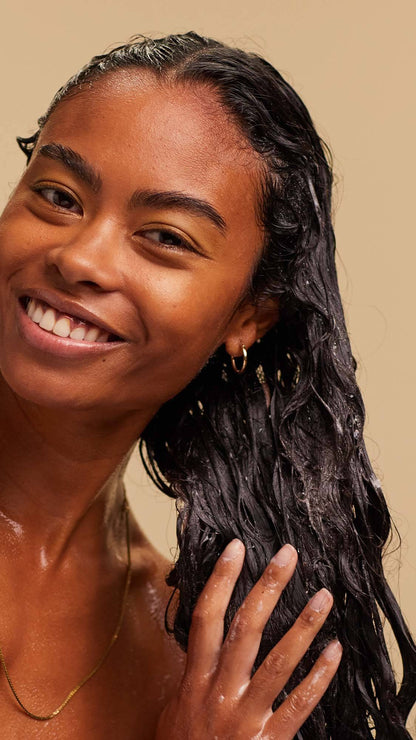 A blissed out, smiling person with long, dark, textured hair massages a white foaming shampoo into the lengths of their hair.