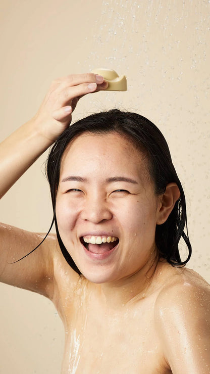 Smiling person using Lush Beirut King of Skin solid body butter on their head under running water, enjoying a refreshing, moisturizing experience.