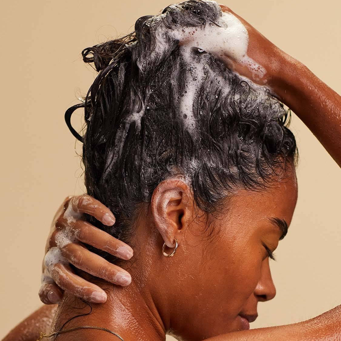 A blissed out, smiling person with long, dark, textured hair massages a white foaming shampoo into the lengths of their hair.