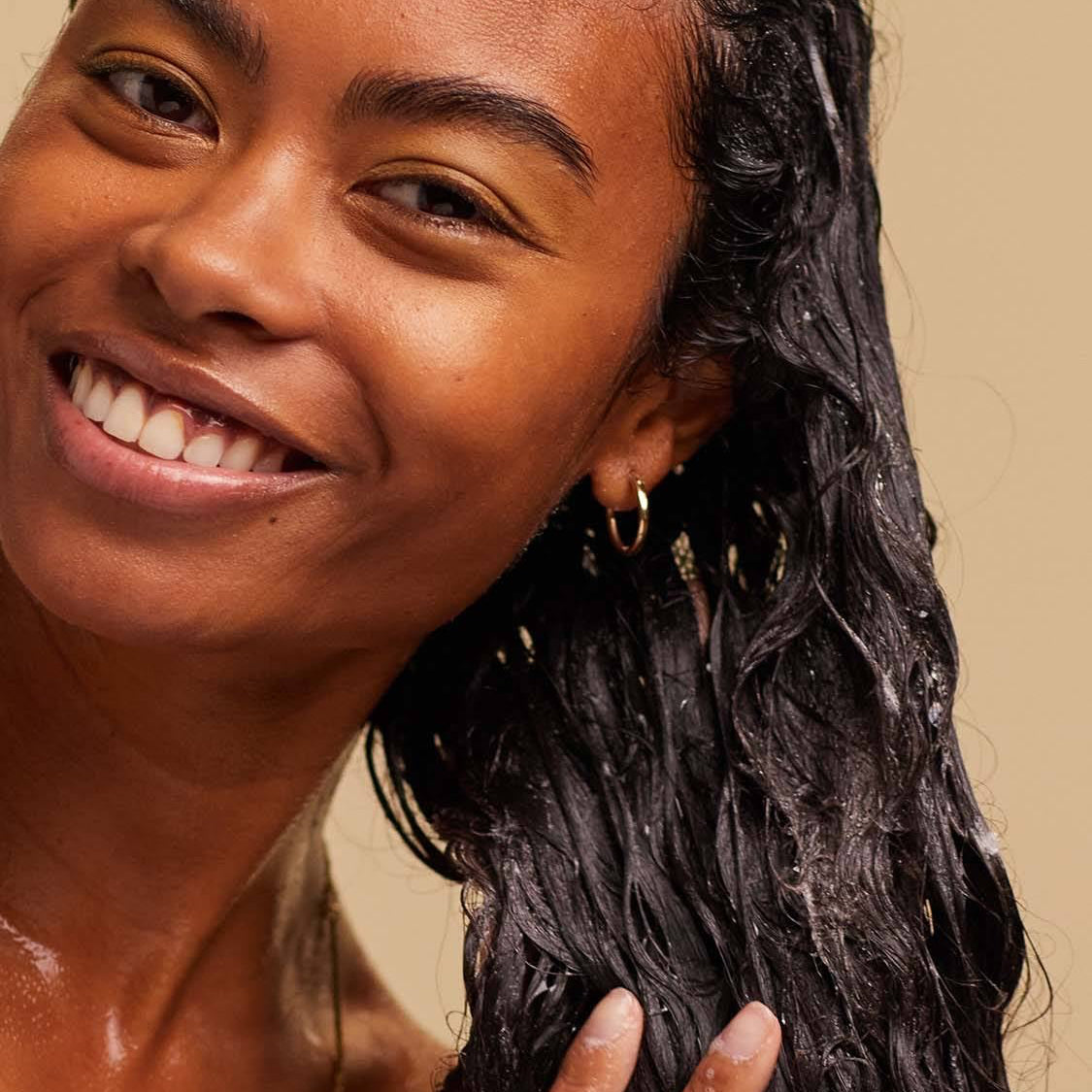 A blissed out, smiling person with long, dark, textured hair massages a white foaming shampoo into the lengths of their hair.