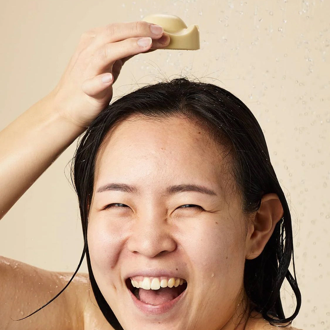 Smiling person using Lush Beirut King of Skin solid body butter on their head under running water, enjoying a refreshing, moisturizing experience.