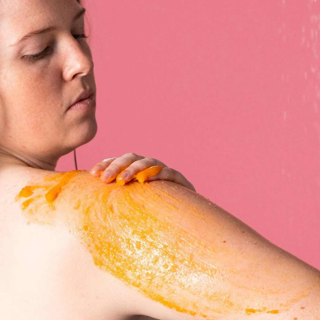 A person with long, wet hair looks down as they rub their arm, which is coated in a bright orange, sea salt body scrub.
